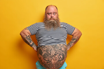 Confident serious blue eyed man with beard, has big abdomen, leads unhealthy lifestyle, dressed in striped undersized sailor t shirt, poses over yellow background. Plump guy stands self assured indoor