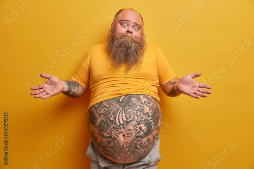 Hesitant thick man with big tattooed belly, shrugs shoulders and looks confused, faces dilemma, makes serious decision, wears undersized yellow t shirt, poses indoor. People and doubt concept