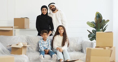 Wall Mural - Portrait of happy muslim family smiling to camera and posing in living room. Moving in new accommodation. Arabian mother, father, daughter and son among carton boxes at home.