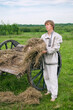 young man in national costume stands near an old hay cart and holds an armful of dry grass in his hands