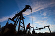 © anatoliy_gleb - Silhouette of engineer in work vest and helmet working on petroleum pump jack. Oil worker using oil pump rocker-machine in oil field under evening sky. Concept of oil extraction and petroleum industry