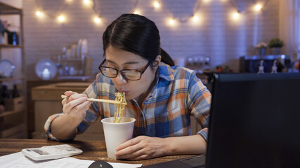 Busy businesswoman eating fast food instant noodle at home kitchen. starving woman enjoy ramen soup sitting at table with laptop. korean lady in glasses holding chopsticks having late night meal