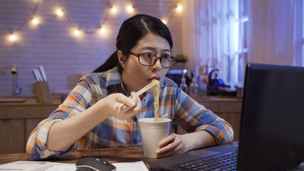 young japanese girl eats instant noodles and looks in laptop computer monitor. serious starving office lady worker working at home in night kitchen and having bedtime snack with unhealthy fast food