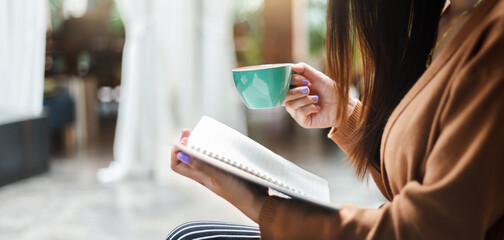 young asian woman holding coffee cup and reading an appointment in book while she relax after work