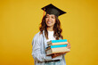 © maxbelchenko - Graduate girl in a graduation hat on her head, with books stands on a yellow background. African American girl posing with smile. Graduation, university, college, distance education concept.