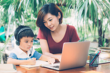 Wall Mural - Young asian mother and son using laptop computer for study and learning together at home, boy writing on notebook for homework and wearing headphone, teacher or mom support child, education concept.
