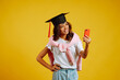 Portrait of African American girl in a graduation hat on her head posing with phone on a yellow background. Graduation, university, college, distance education concept.
