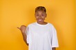 © Roquillo - Lovely Young African American girl with afro short hair wearing white tshirt standing over isolated yellow wall pointing aside with forefinger, showing at copy space having news about bargains