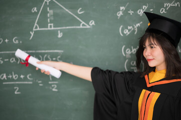 Wall Mural - Asian female graduates wear a black robe and black hat with golden tassels. Standing in front of the blackboard in the classroom