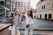 Attractive Blonde Girl In White Pants Posing In City Center. Outdoor Photo Of Gorgeous Female Friends Dancing In Square.