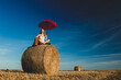 blonde girl with suitcase and umbrella is sitting on a rolled haystack in field in sunset time