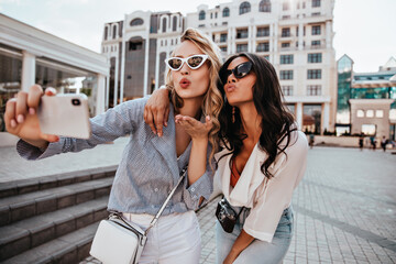 Wall Mural - Happy blonde girl in white sunglasses making selfie with kissing face expression. Glamorous female friends expressing love for photo.