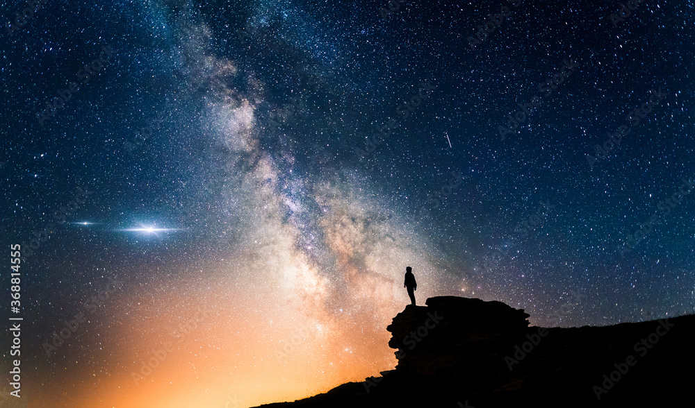 Man standing in a high place looking up in wonder to the Milky Way galaxy. Small silhouette of a man under the Milky Way and the magical starry sky. Concept of human smallness. - obrazy, fototapety, plakaty 