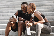 After Training. Happy African Couple Resting On Stairs Outdoors And Using Smartphone
