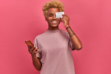 Happy Young Dark Skinned Freelancer Woman Holding Smartphone And Bank Card, Which Closes One Eye On Pink Background. Girl Is Happy That Now She Will Receive Good Salary On Card. Lifestyle And Carefree
