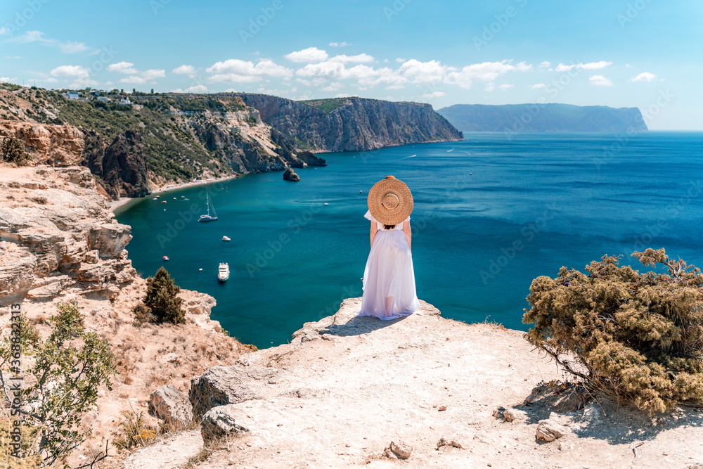 From behind, a woman is seen in a white flying dress fluttering in the wind. In a straw hat, walking down the stairs against the background of the sea with rocks and boats. The concept of travel - obrazy, fototapety, plakaty 