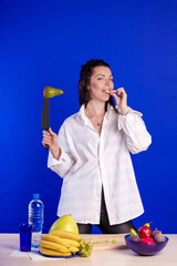 Emotional actress woman in a white shirt posing with fruits and a knife in her hands and on the table on a blue background