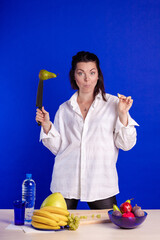 Emotional actress woman in a white shirt posing with fruits and a knife in her hands and on the table on a blue background