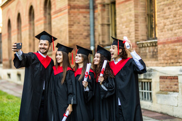 Wall Mural - Capturing a happy moment. Four college graduates in graduation gowns standing close to each other and making selfie