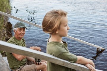 Wall Mural - Mature father with his son outdoors sitting on wooden setting by lake or river, spending time near water in summer day, wearing green t shirts, enjoying having rest together.