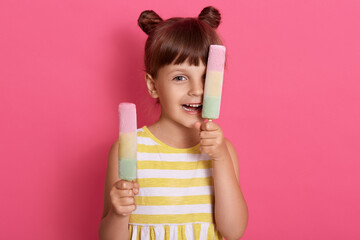 Little Caucasian girl kid expressing happiness, holds up two sorbets and covering her eye with one fruit lollies, standing against rose wall, having fun in summer.