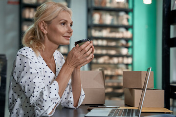 Wall Mural - Enjoying morning coffee. Side view of a beautiful and happy female business owner or business woman drinking coffee while working in her art studio or craft pottery shop