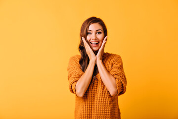 Wall Mural - Ecstatic beautiful woman touching her face. Studio portrait of smiling carefree girl isolated on yellow background.