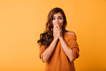 Wall Mural - Shocked beautiful girl in orange attire covering face with hands. Indoor photo of charming white lady posing on yellow background with amazement.