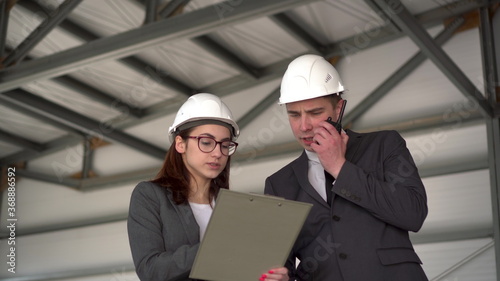 Young man and woman in helmets with documents at a construction site. Bosses in suits are discussing an architecture project. A man speaks on a walkie-talkie. People look around.