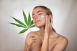 Woman applying CBD facial cream made from cannabis extract for a natural skin treatment. Portrait of young woman with cannabis leaf. Cosmetology and treatment concept. Isolated on gray background