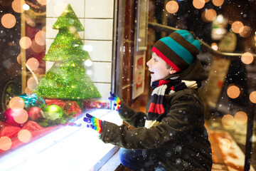 Wall Mural - Cute little healthy school kid boy on Christmas market. Funny happy child in fashion winter clothes making window shopping decorated with gifts, xmas tree. Snow falling down, snowfall