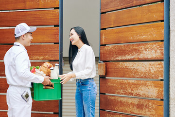 Wall Mural - Smiling courier deliverying milk and fresh groceries to pretty happy young woman