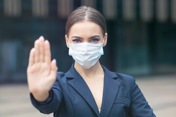 Beautiful girl, young woman in protective medical mask on her face outdoors, serious businesswoman showing palm, hand, stop no sign against covid-19. Virus, pandemic, epidemic coronavirus concept.