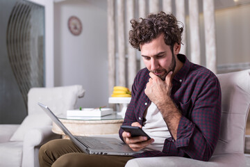 Wall Mural - Young handsome man making video call with friends while sitting on sofa at his modern home.Concept of happy business people. Business man at home checking statistics on mobile phone and laptop