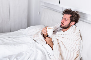 Sick day at home. Handsome Man has fever and common cold. Cough. Closeup Of Young Man Caught Cold Or Flu Illness. Portrait Of Unhealthy man Feeling Pain In Throat and drinking hot beverage.