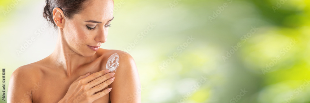 Beautiful young woman applying cream to her shoulder. Skincare spa wellness and cosmetics concept. Blurred green panoramic banner - obrazy, fototapety, plakaty 