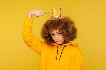 Look, I am best! Selfish haughty curly-haired woman pointing at crown on head and looking with arrogance supercilious, being egoistic with over-inflated ego. studio shot isolated on yellow background