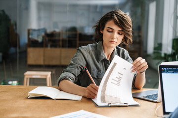 Image of young focused woman writing down notes while sitting at table