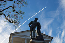 The Statue Of Hercules In Front Of The Cameron Gallery In Catherine Park Against A Background Of Blue Sky, A Tree And A High-flying Plane.