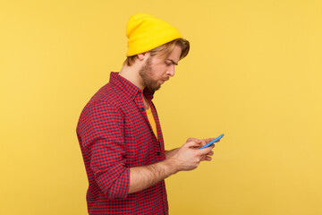 Wall Mural - side view trendy guy in hipster beanie hat and shirt using cell phone, texting or reading message, c