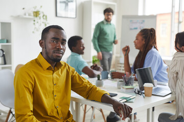 Wall Mural - Portrait of contemporary African-American man looking at camera while sitting at table during meeting with business team in background, copy space