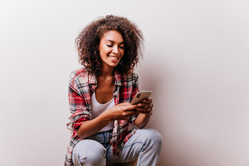 Wall Mural - Blithesome black woman in checkered shirt using her smartphone. Appealing girl reading phone message.