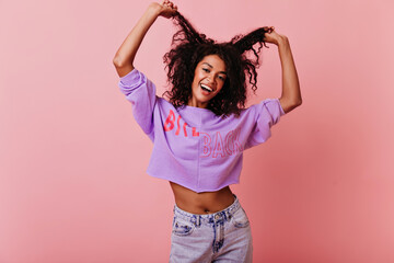 Wall Mural - Sensual black girl posing on rosy background with happy face expression. Jocund african woman having fun in studio with pink interior.