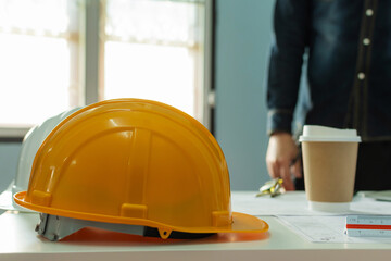 Wall Mural - yellow safety helmet and coffee cup on workplace desk with architect or engineer standing in meeting room office center at construction site, contractor meeting, engineering and construction concept