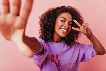 Wall Mural - Appealing african woman making selfie with peace sign. Indoor photo of emotional laughing girl posing on pink background.