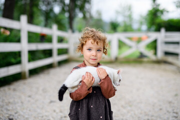 Wall Mural - Small girl with cat standing on farm, looking at camera.