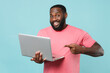 © ViDi Studio - Smiling young african american man guy in casual pink t-shirt isolated on blue background studio portrait. People lifestyle concept. Mock up copy space. Pointing index finger on laptop pc computer.
