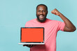 © ViDi Studio - Happy young african american man in casual pink t-shirt isolated on blue background. People lifestyle concept. Mock up copy space. Hold laptop pc computer with blank empty screen doing winner gesture.