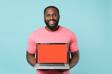 Smiling young african american man in casual pink t-shirt isolated on blue background studio portrait. People lifestyle concept. Mock up copy space. Hold laptop pc computer with blank empty screen.
