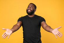 Smiling African American Man Guy Football Fan In Casual Black T-shirt Isolated On Yellow Background Studio. People Lifestyle Concept. Mock Up Copy Space. Standing With Outstretched Hands For Hugs.
